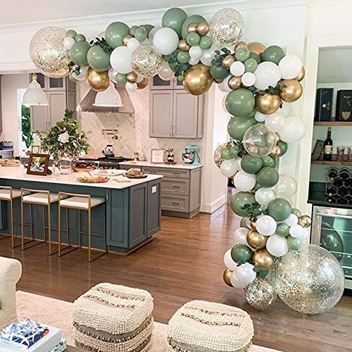 A decorative balloon garland arch kit with a variety of balloons in olive green, white, and gold colors, assembled over a room setting with chairs and a table.