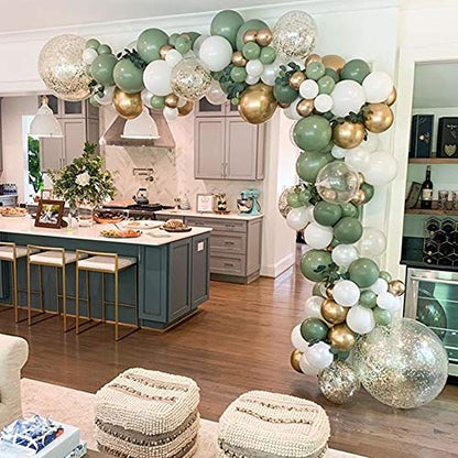 A decorative balloon garland arch kit with a variety of balloons in olive green, white, and gold colors, assembled over a room setting with chairs and a table.