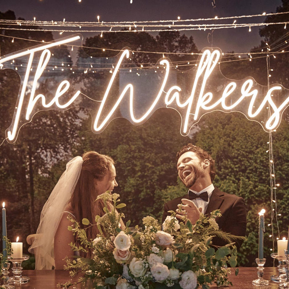 Neon sign reading 'The Walkers' above a couple at a wedding table with floral arrangements and candles.